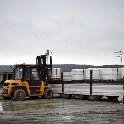 A forklift is loading cargo onto a truck in a muddy logistics area with a worker assisting.